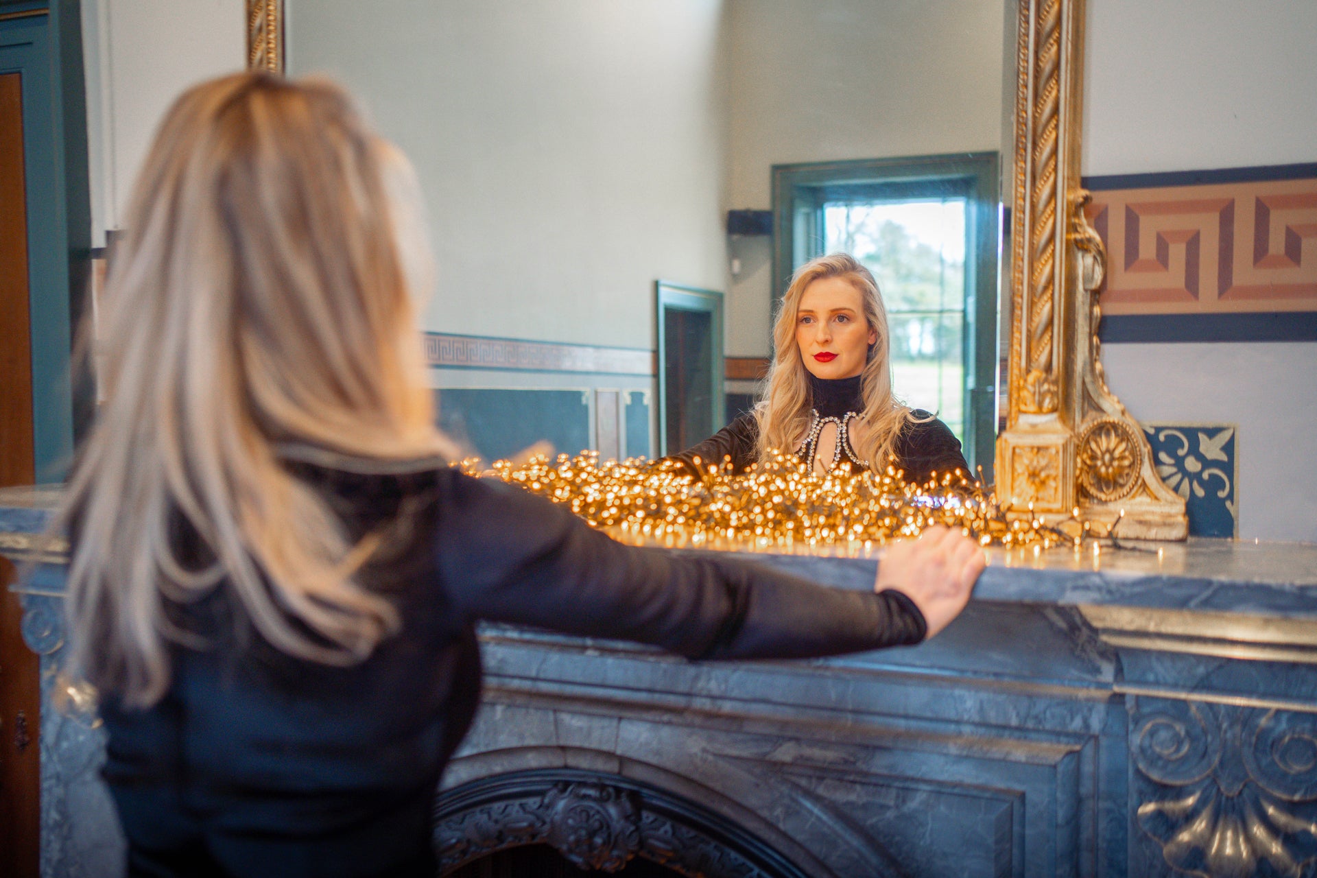 Blonde woman with red lipstick wearing a black dress looking in the mirror